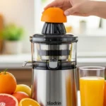 Extractor de jugo con una mano colocando una naranja en la parte superior, rodeado de frutas y un vaso de jugo.